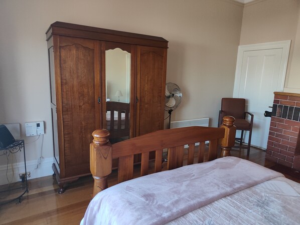 A bedroom with a wooden wardrobe, a bed with a white bedspread, a chair, a fan, and a radiator. - Hawthorn House Launceston (Invermay)