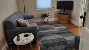 A living room with a grey sofa, a small white side table, a black TV on a wooden stand, and a patterned rug. - Hawthorn House Launceston (Invermay)