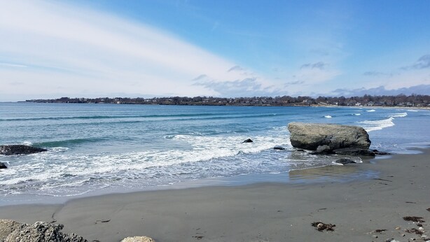 A beach with waves, a large rock, and a distant shoreline with buildings. - Immaculate Cottage, Huge Deck And Great Back Yard For All Your Yard Games. (Middletown)
