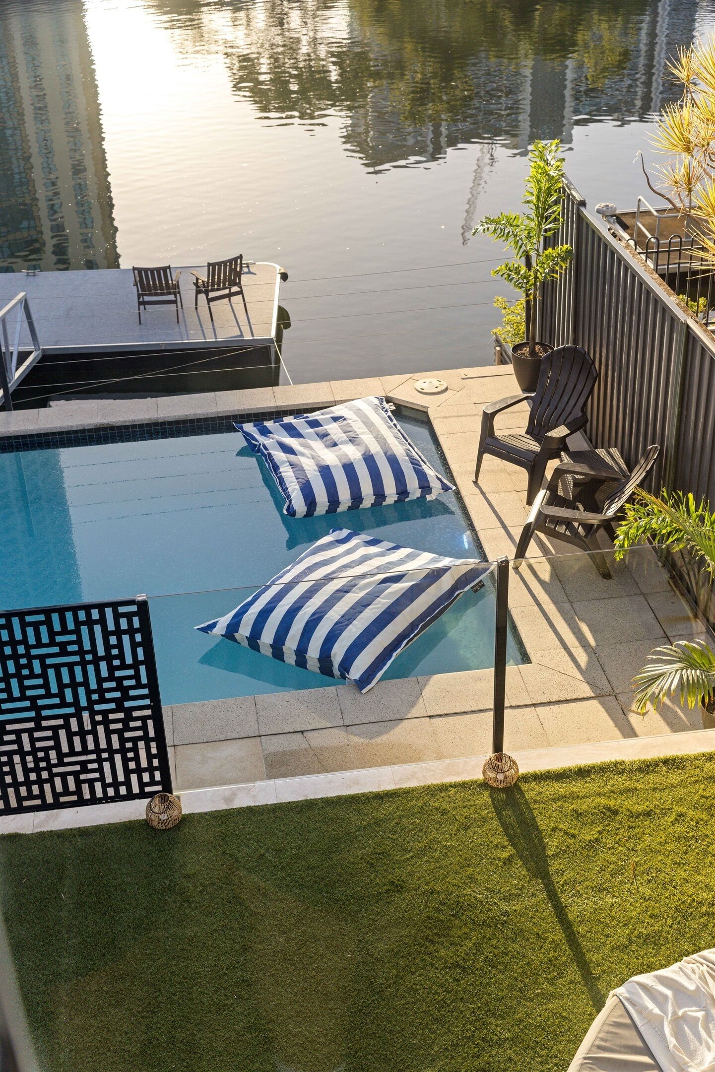 A swimming pool with lounge chairs and striped pillows, surrounded by a fence and greenery.