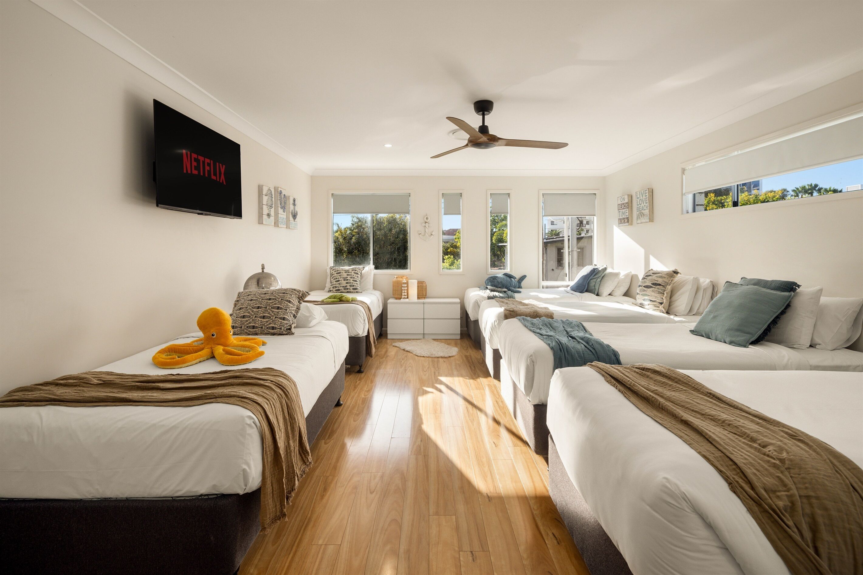 A hotel room with three beds, a ceiling fan, a TV on the wall, and large windows.