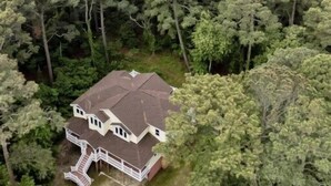 Una casa de dos pisos con un porche que rodea toda la estructura, rodeada de un bosque frondoso y una playa al fondo.