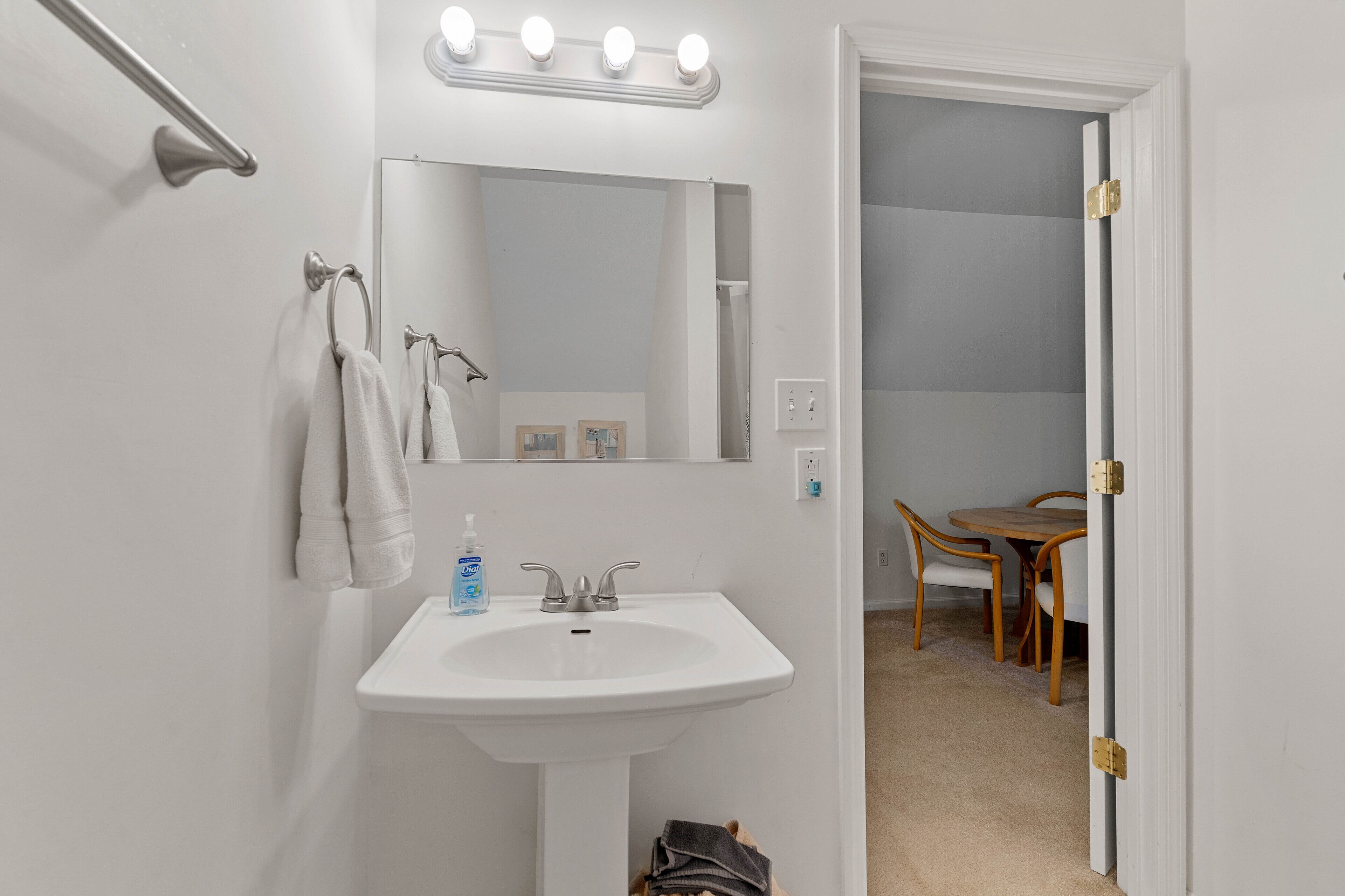 Une salle de bain avec un lavabo blanc, un miroir avec éclairage intégré et une porte donnant sur un coin repas.
