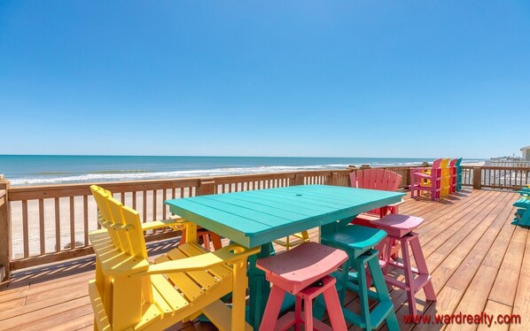 Eine Holzterrasse mit einem türkisfarbenen Tisch und bunten Adirondack-Stühlen mit Blick auf den Strand.
