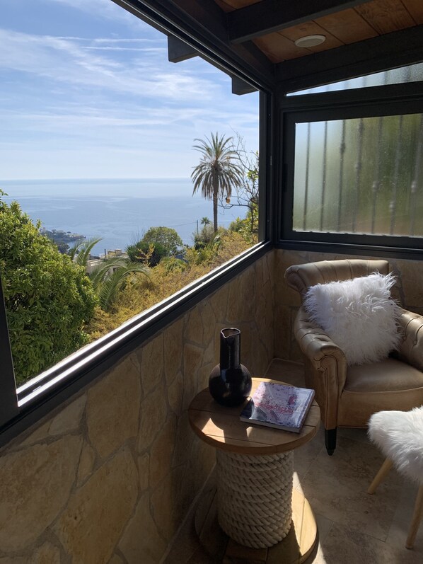 A balcony with a view of the ocean, a stone wall, a leather chair with a fluffy pillow, a small round table with a vase and a magazine, and a woven basket.