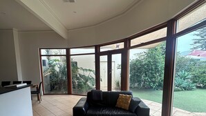 A spacious living room with a black leather sofa, a white coffee table, and large sliding glass doors leading to a garden. - Esplanade Beach House with beautiful sea views (Semaphore)