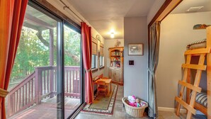 A cozy room with a wooden bunk bed, a dining table with chairs, a large window with curtains, and a bookshelf.