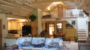 A spacious living room with a high ceiling and wooden beams, a dining area with a table and chairs, and a kitchen with wooden cabinets and appliances.