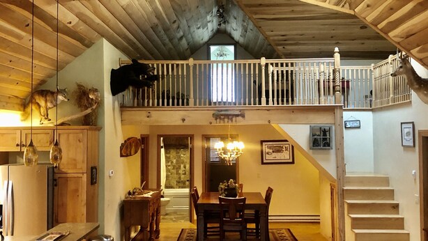 A spacious dining area with a high ceiling and wooden beams, a balcony with a railing, and a staircase leading to an upper level.