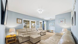 A living room with a beige sectional sofa, a wooden coffee table, and a small side table with a lamp. There are framed pictures on the walls and a window with curtains.