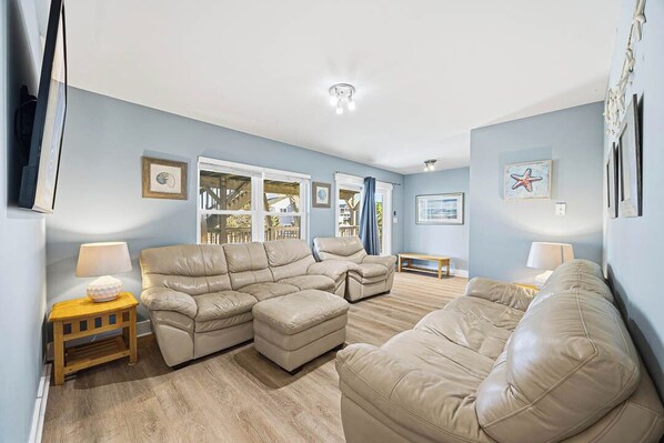 A living room with a beige sectional sofa, a wooden coffee table, and a small side table with a lamp. There are framed pictures on the walls and a window with curtains. - Cabana Royale | 520 ft from the beach | Private Pool (Virginia Beach)