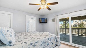 Quarto com cama, ventilador de teto, televisão e porta de vidro deslizante para um terraço.