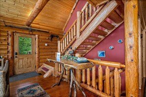 A log cabin interior with a wooden staircase, a wooden table with a book and decorative items, and a door with a view of greenery outside.