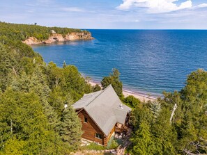 A cabin with a sloped roof, surrounded by dense forest, overlooking a calm body of water and a distant rocky coastline.