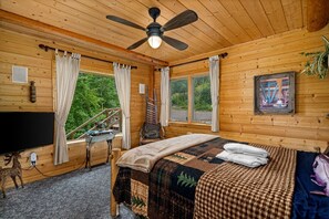 Ein Schlafzimmer einer Holzhütte mit einem Bett, einem Deckenventilator, einem Fernseher und einem Fenster mit Blick auf einen Wald.