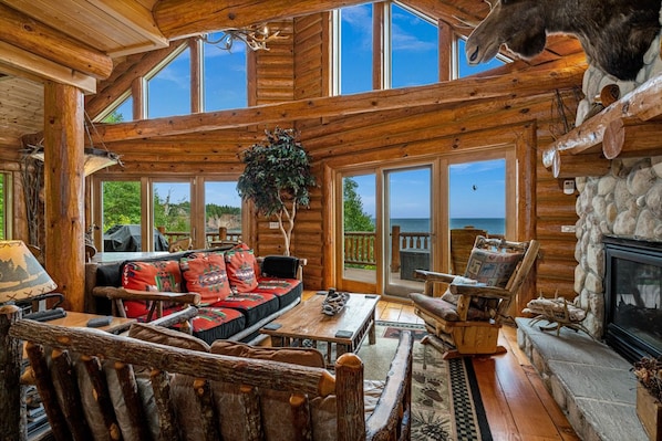 A log cabin interior with a fireplace, wooden furniture, and large windows.