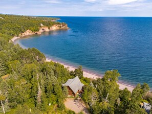 A cabin surrounded by dense forest, overlooking a curved coastline with a beach.