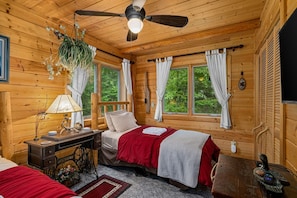 A wooden cabin bedroom with a bed, a desk, a ceiling fan, and a window with curtains.