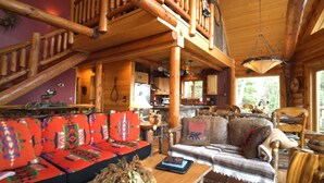 A rustic cabin interior with a wooden staircase, a sofa with colorful cushions, a dining table, and a kitchen area. - Lake Superior Luxury Log Home, Sauna, Waterfront, Amazing Views (Two Harbors)