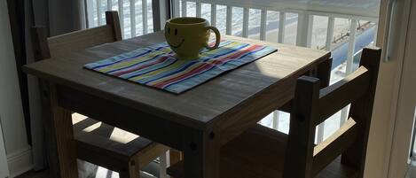 A wooden table with a yellow mug on a colorful placemat, overlooking a beach with waves.