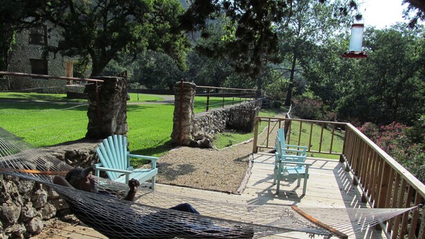 A hammock on a wooden deck with two light blue Adirondack chairs, a stone wall, and a grassy yard with trees. - SPECTACULAR LODGE ON PRIVATE RANCH. PERFECT FOR SMALL AND LARGE GATHERINGS! (LEAKEY)