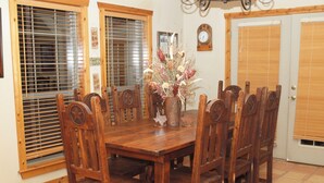 A wooden dining table with eight chairs, surrounded by windows with blinds, a clock on the wall, and a door with a wooden frame.
