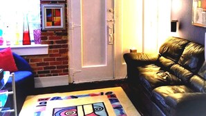 A living room with a brick wall, a white door, a black leather sofa, and a colorful rug. - The Gallery Getaway in the Heart of Historic, Riverfront Wilmington!  (Wilmington)