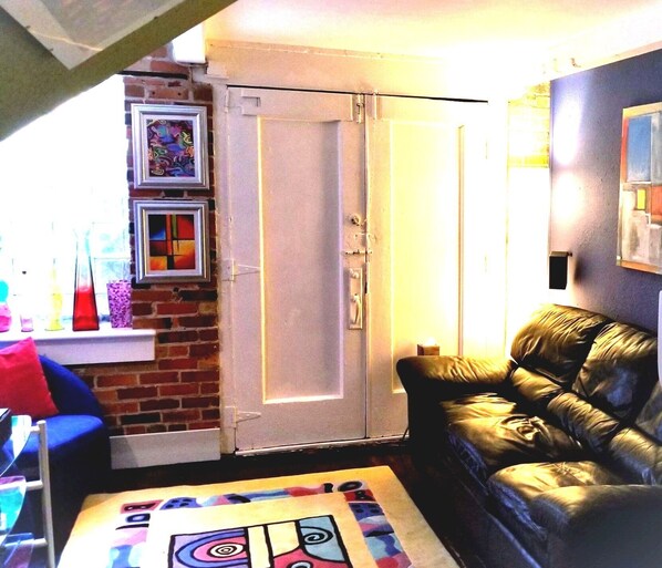 Uma sala de estar com parede de tijolos, porta branca, sofá de couro preto e um tapete colorido.