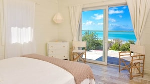 A bedroom with a bed, a dresser, two chairs, and a large sliding glass door leading to a balcony with a view of the beach. - Sueno De Mar: Beachfront Villa for 10 (Hartswell)