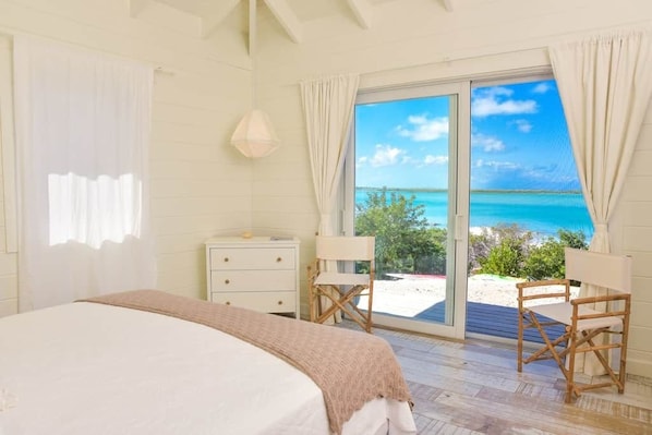 A bedroom with a bed, a dresser, two chairs, and a large sliding glass door leading to a balcony with a view of the beach.