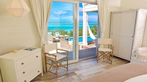 A bedroom with a bed, a white dresser, two wooden chairs, a white nightstand, and a view of the ocean through a sliding glass door. - Sueno De Mar: Beachfront Villa for 10 (Hartswell)