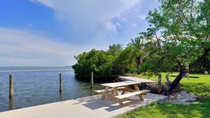 A wooden dock extending into a calm body of water, with a picnic table and lush greenery nearby. - Private Oceanfront Cottage with dockage, pool just minutes to Key West! (Cudjoe Key)