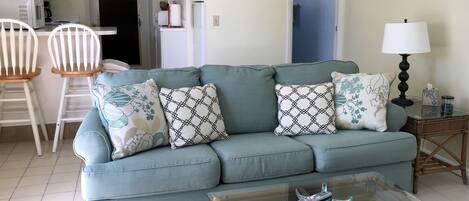 A living room with a light blue sofa, two patterned pillows, a glass coffee table with a remote control, a lamp, and a small table with a tissue box.
