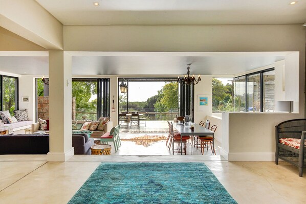 A spacious living area with a large blue rug, a dining area with a table and chairs, and a view of the outdoors through large windows. - Solar-Powered Table Mountain Retreat, Kid-Friendly Space with Lush Garden & Pool (Cape Town)