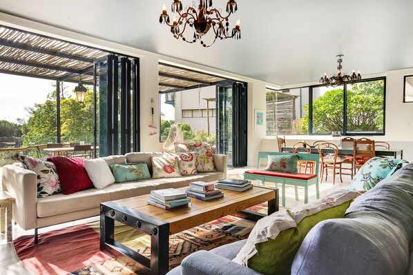 A spacious living area with a sofa, coffee table, and dining table, featuring large windows and a chandelier.