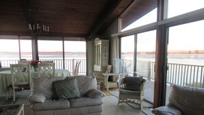 A spacious room with a large window overlooking a body of water, a white couch, wicker chairs, and a dining area with a table and chairs. - The Waterfront Deckhouse-Pine Point Beach Waterfront Property (Scarborough)
