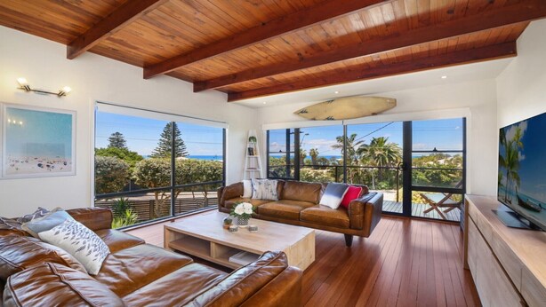 A spacious living room with a wooden ceiling, large windows offering a view of palm trees and the ocean, a leather sofa, and a television on the right. - MYSTIC WAVES | WIFI , Pet Friendly | Accom Holidays (WAMBERAL)
