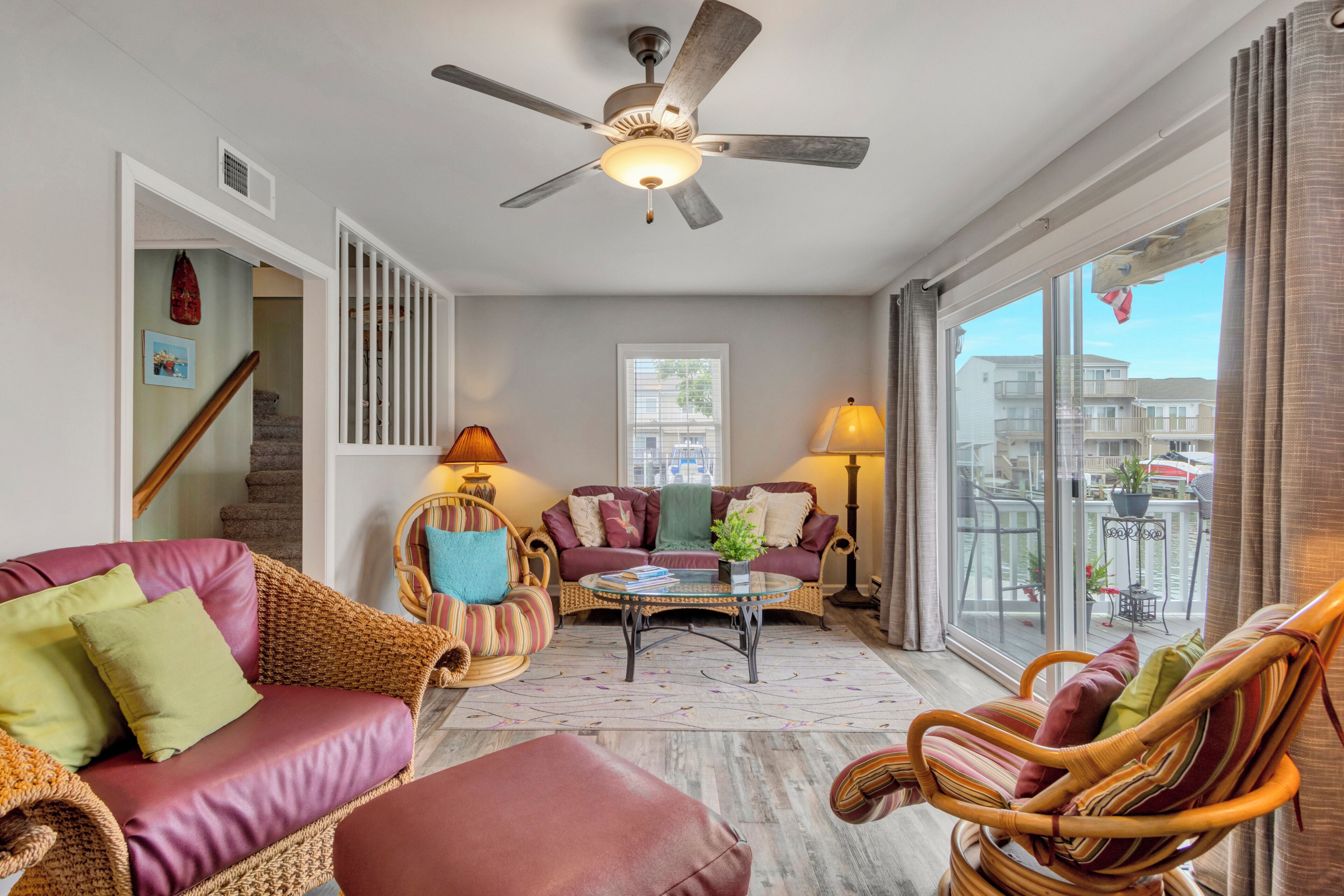 A spacious living room with a ceiling fan, wicker furniture, and a large window offering a view of the outside.