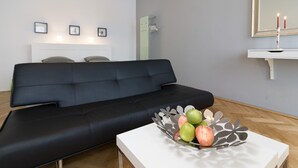 Un salon moderne avec un canapé noir, une table basse blanche agrémentée d’une composition décorative de pommes, un miroir mural et une petite étagère blanche sur laquelle est posée une bougie.