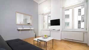 Un salon moderne avec un canapé noir, une table basse blanche sur laquelle se trouve un panier de fruits, un téléviseur à écran plat fixé au mur et de grandes fenêtres équipées de stores.