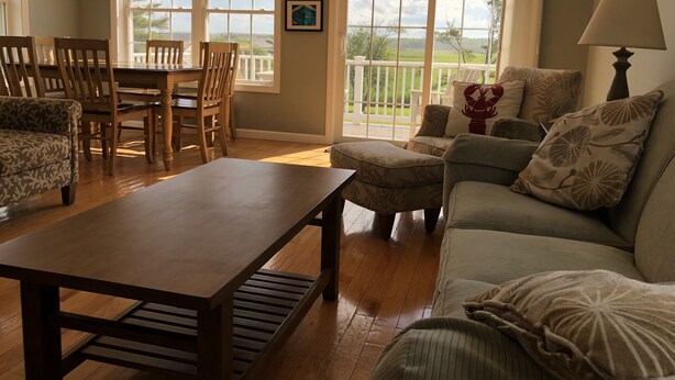 A spacious living room with a wooden coffee table, a sofa, and a dining area with chairs and a table. Large windows provide views of the outdoors. - Lovely house with gorgeous marsh views! (Wells)