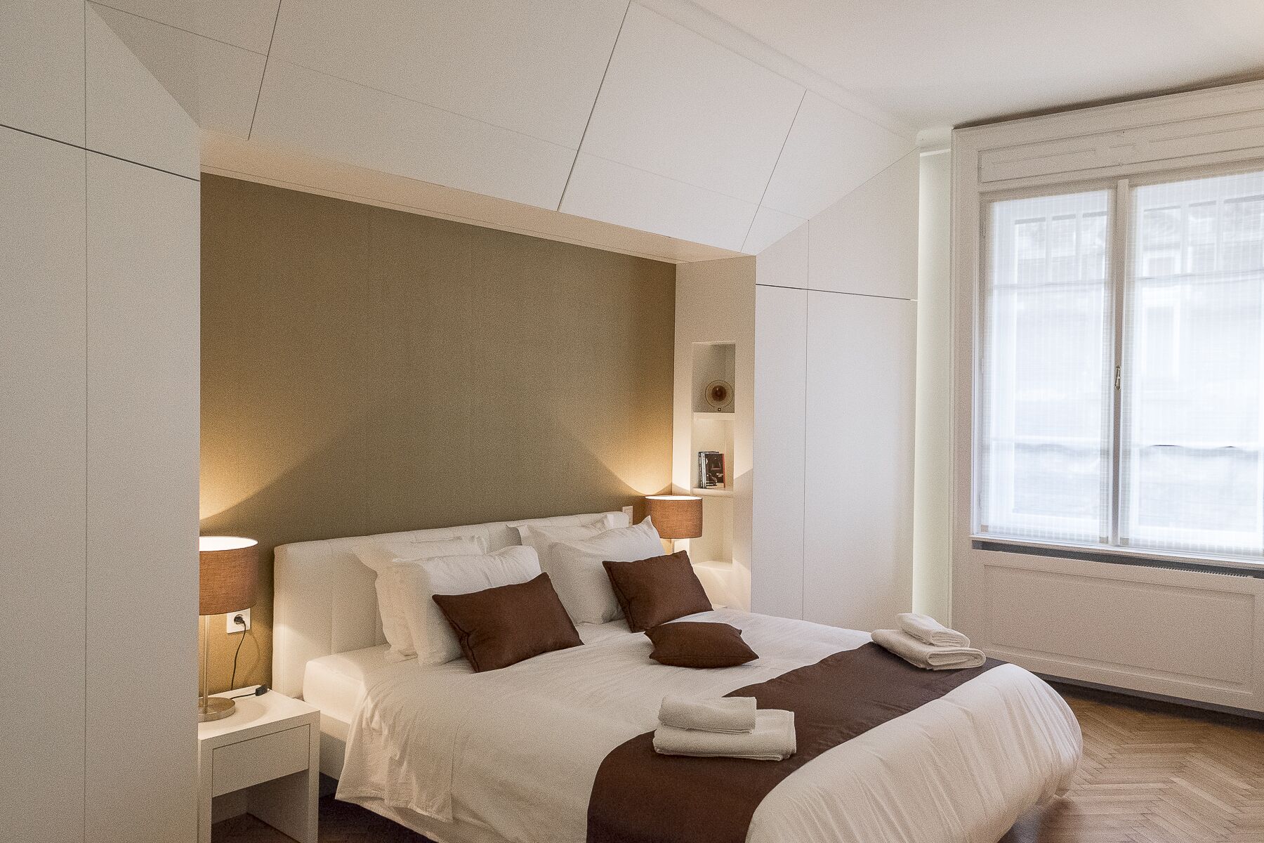 Ein Hotelzimmer mit einem großen Bett, Nachttischlampen, einem Nachttisch, einem Fenster mit Jalousien und einem an der Wand befestigten Regal mit Büchern.