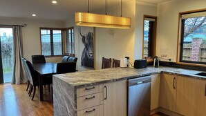 A modern kitchen with a marble countertop, wooden cabinets, and a built-in dishwasher. There is a dining area with a table and chairs, and a large window with blinds.