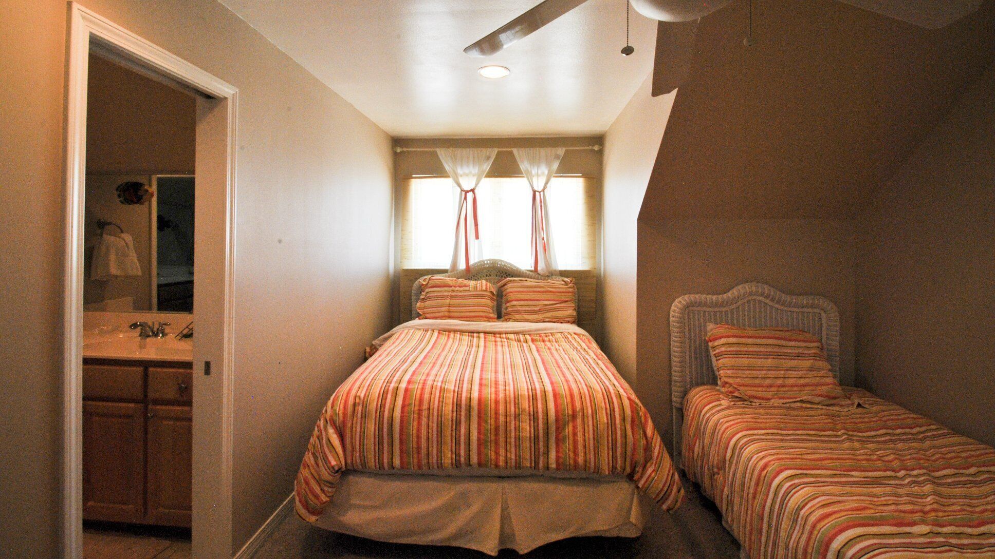 A bedroom with a single bed, a smaller bed, a window with curtains, a ceiling fan, and a bathroom with a sink and mirror.