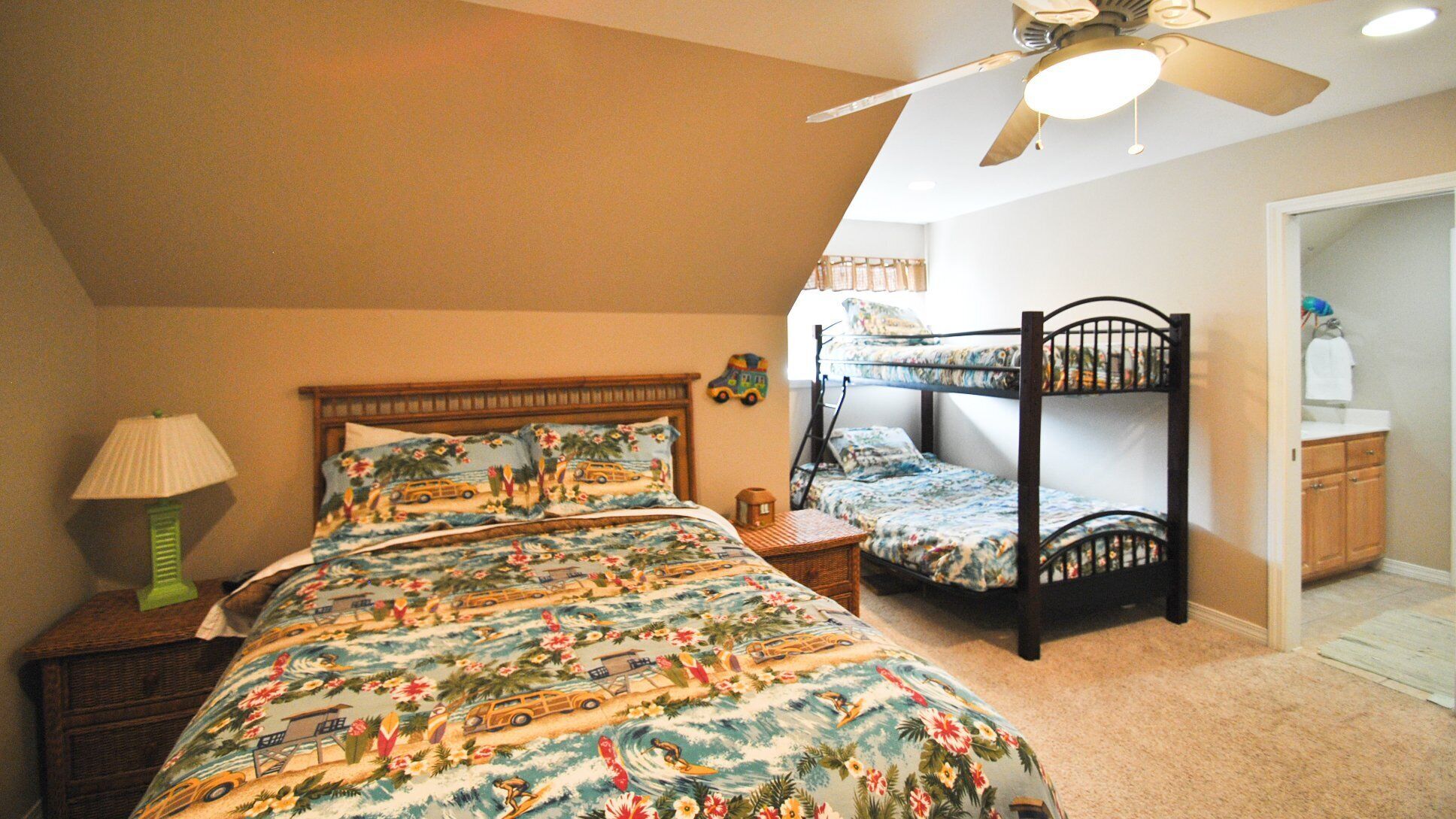 A bedroom with a bunk bed, a single bed with floral bedding, a nightstand, a lamp, and a ceiling fan.