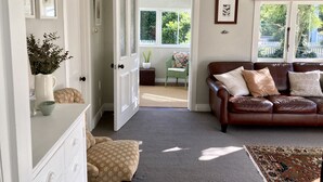 A living room with a brown leather sofa, a patterned armchair, a coffee table, and a rug. There is a door leading to an outdoor area with a chair and a plant.
