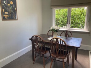 A dining room with a wooden table and chairs, a vase with flowers, and a window with a view of greenery.