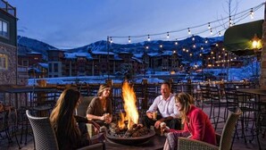 Um grupo de pessoas sentadas ao redor de uma fogueira em um pátio externo, com luzes de corda acima, cercado por edifícios e montanhas cobertas de neve ao fundo.