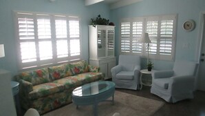 Una sala de estar con un sofá floral, dos sillones blancos, una mesa de centro de vidrio y un aparador con una planta encima.