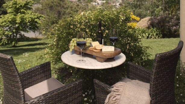 Una mesa de mimbre con una tabla de quesos, uvas y copas de vino, dispuesta al aire libre.